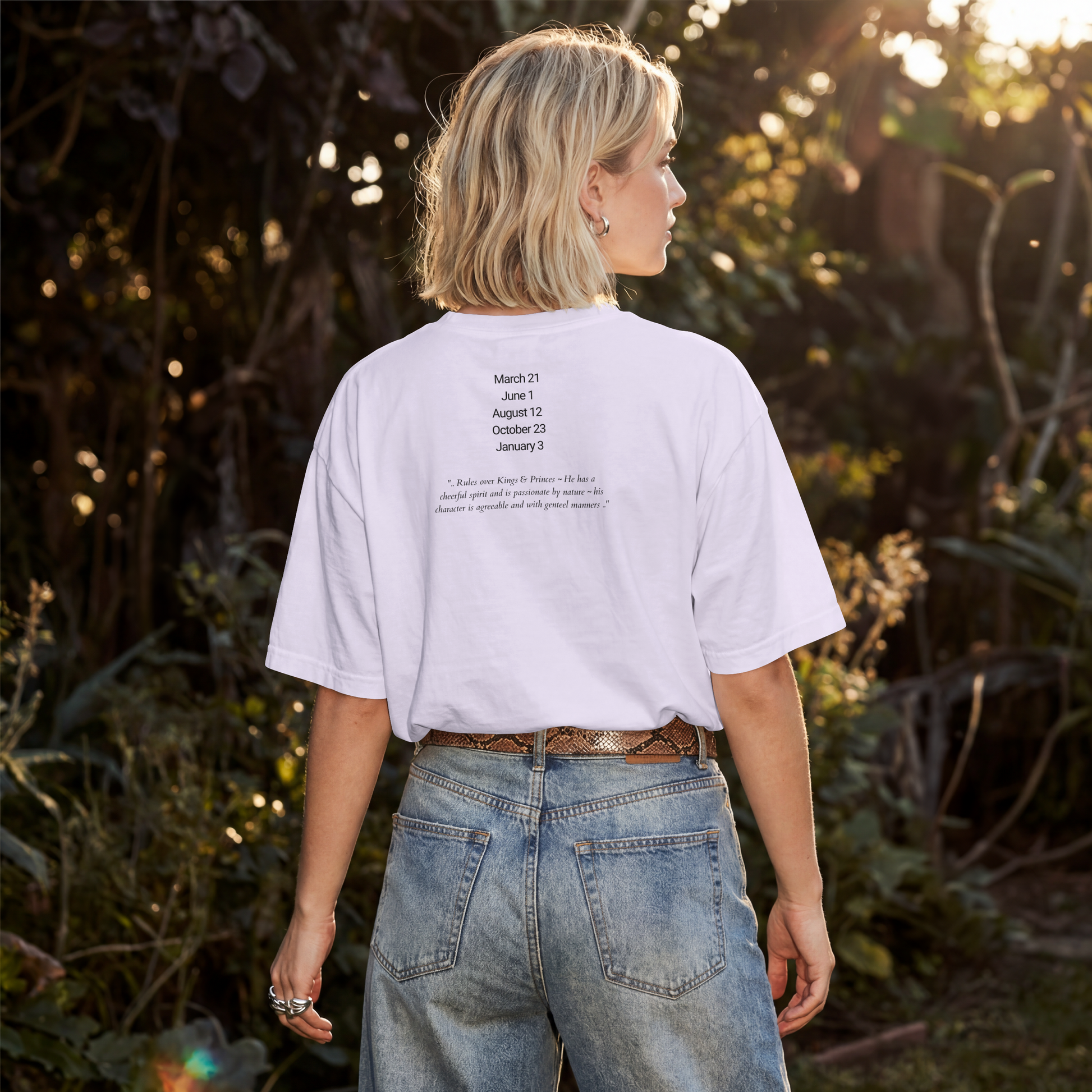 A woman viewed from behind wearing a white graphic t-shirt with black text listing dates and a quote, paired with light-wash blue jeans, set against a sunlit, blurred outdoor background of foliage.